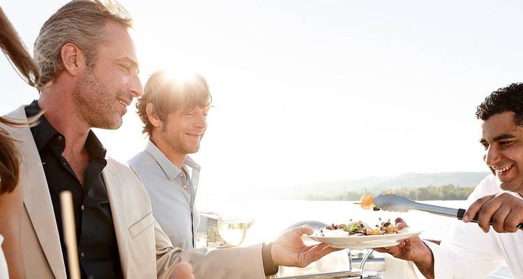 Des invités sur un pont de croisière baigné de soleil souriaient tandis qu'un chef servait un plat gastronomique avec un arrière-plan pittoresque.