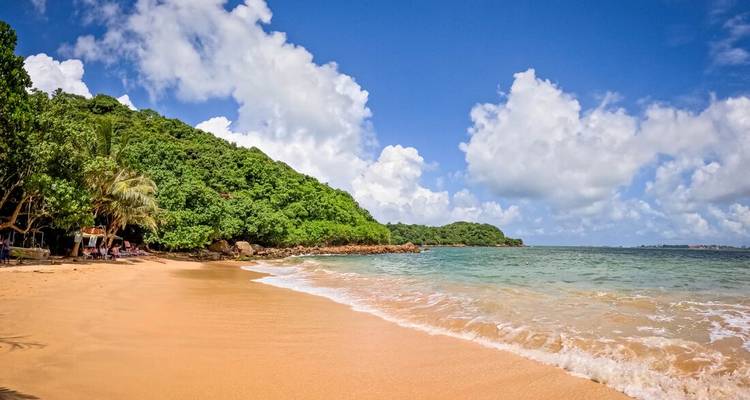 Plage tropicale immaculée avec du sable doré, un promontoire verdoyant et un océan turquoise sous des nuages cotonneux.