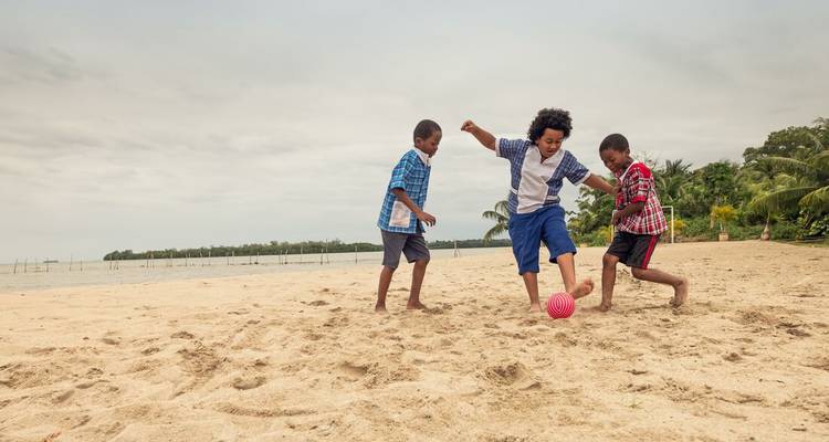 Drei Kinder spielen energisch Fußball an einem sandigen Strand unter einem grauen Himmel.