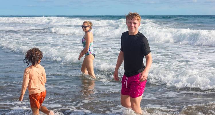 Drei Kinder spielen in der seichten Brandung eines Sandstrandes mit rollenden Wellen hinter ihnen.
