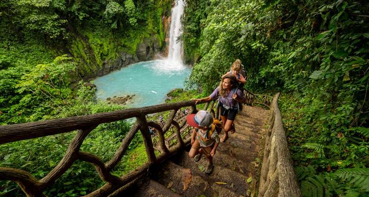 Une famille gravit des escaliers de pierre à travers la forêt tropicale vers une cascade turquoise qui plonge dans un bassin en contrebas.
