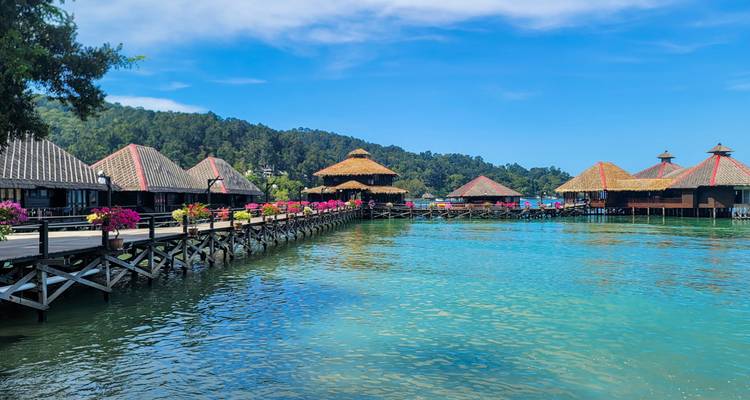 Des bungalows en bois sur pilotis bordent un lagon turquoise adossé à des collines boisées.