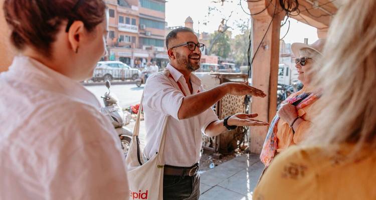 Un guide local explique avec animation un site à deux voyageurs dans une rue indienne animée.