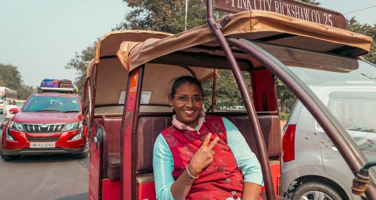 Une femme souriante fait un signe de paix assise dans un pousse-pousse électrique rouge.