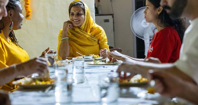 Les invités partagent un repas fait maison avec une hôtesse souriante vêtue de jaune à l'intérieur d'une maison indienne.