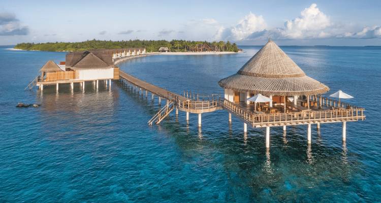 Une vue aérienne panoramique d'une jetée en bois courbée menant à des villas au toit de chaume au-dessus des eaux turquoise maldiviennes.