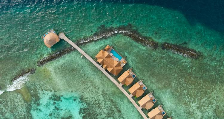 Vue aérienne de villas sur pilotis avec piscines privées s'étendant le long d'une jetée au-dessus d'une mer parsemée de récifs.