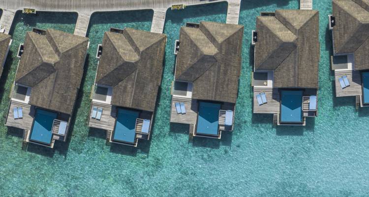 Plan de drone symétrique de bungalows au sol vitré flottant au-dessus d'une eau bleue scintillante.