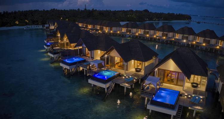 Vue nocturne de villas de luxe sur l'eau qui brillent au bord d'un lagon turquoise sous un ciel sans étoiles.