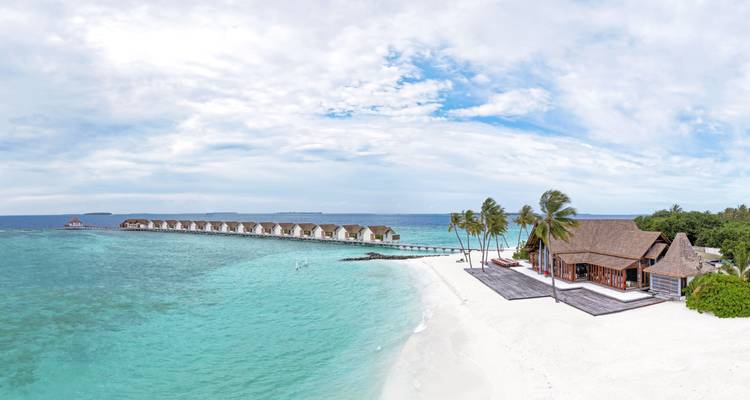 Vue aérienne panoramique d'une plage bordée de palmiers et d'une longue jetée de villas s'étendant dans une mer turquoise.