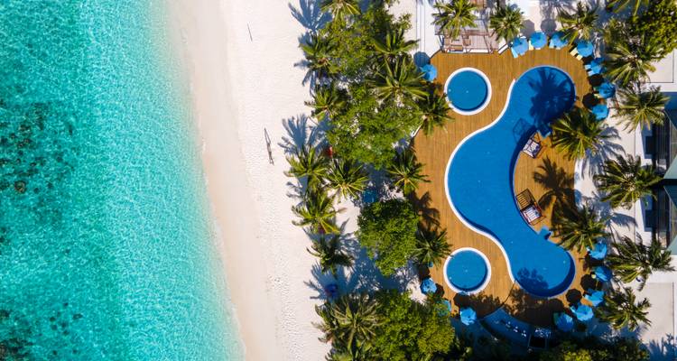 Vue à vol d'oiseau colorée d'une piscine sinueuse de complexe hôtelier encadrée de palmiers près d'un lagon bleu néon et d'une plage.