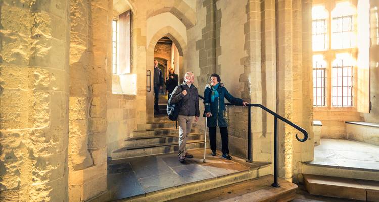Des visiteurs âgés montant des marches en pierre à l'intérieur d'une salle voûtée historique éclairée par une lumière du jour chaleureuse