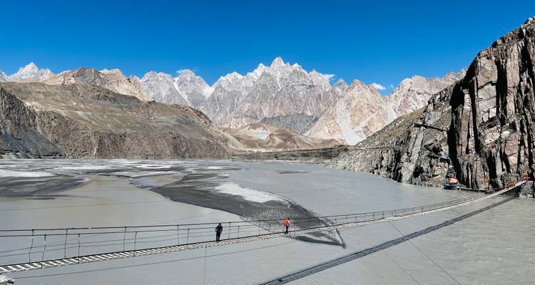 Hikers crossing a narrow suspension bridge above a silty river with the dramatic Passu Cones behind.