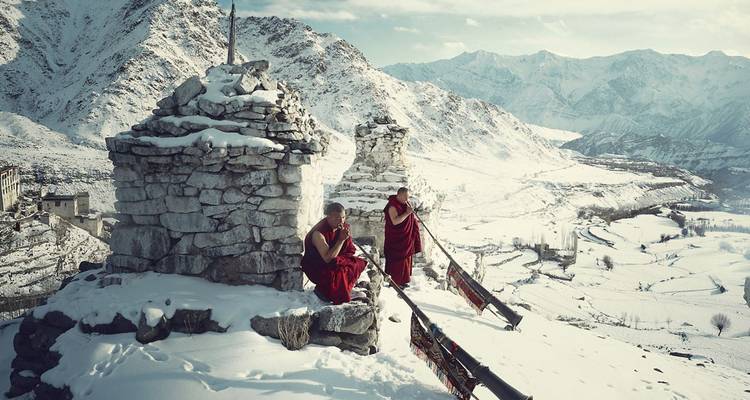 Two monks in red robes meditate beside ancient stone chortens overlooking a snow-blanketed Himalayan valley.