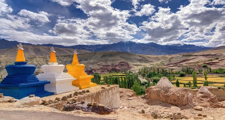 Colorful chortens overlook patchwork fields and distant Himalayan ridges beneath dramatic clouds.