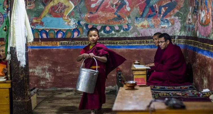 Inside a dimly lit monastery, a novice monk carries a metal pail past elders reading scriptures.