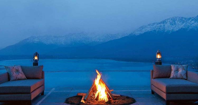 Gemütliche Feuerstelle zwischen zwei Lounge-Sofas mit Blick auf einen ruhigen See und verschneite Berge in der Dämmerung.