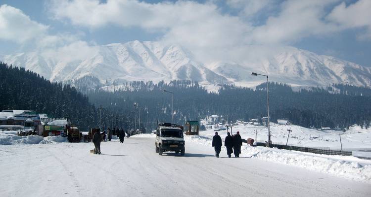 Schneebedeckte Hauptstraße in Gulmarg mit Fahrzeugen und Menschen, umrahmt von Kiefernwäldern und Gipfeln.