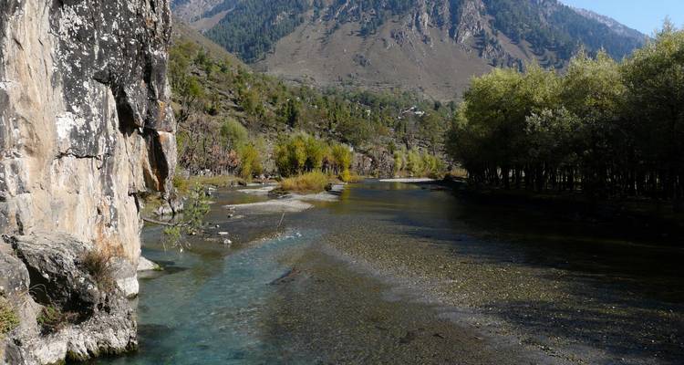 Arroyo sereno de montaña bordeado por un acantilado rocoso y grupos de árboles con altas laderas áridas en el fondo