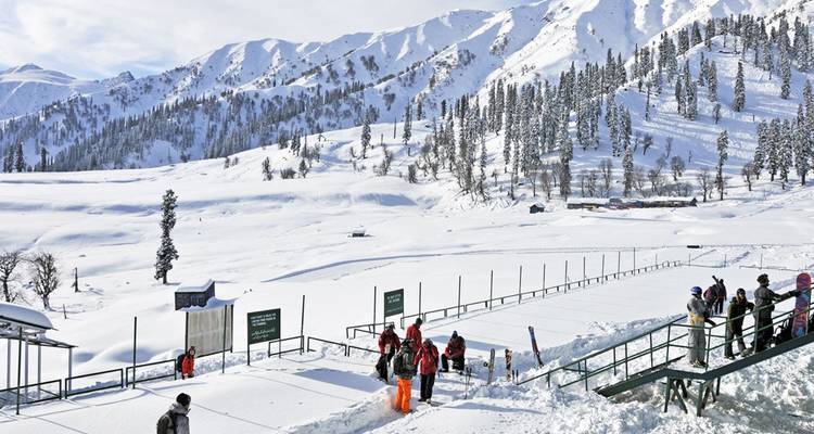 Cuenca alpina cubierta de nieve con esquiadores y snowboarders reunidos cerca de tableros informativos con un fondo de picos blancos bordeados de pinos