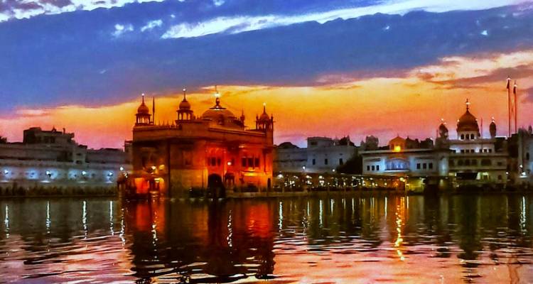 The Golden Temple illuminated at dusk, reflecting vividly in the surrounding holy pool under a colourful sky.