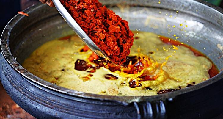 Close-up of vibrant red spices being poured into a bubbling yellow curry in a large metal pan.
