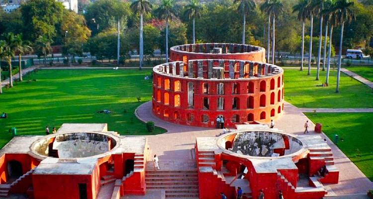 Aerial view of the red-stone astronomical instruments at Jantar Mantar surrounded by green lawns and palm trees.