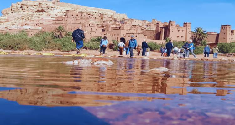 Reisende waten durch einen seichten Fluss auf die Lehmziegelfestung Ait Benhaddou zu, die sich im Wasser spiegelt.