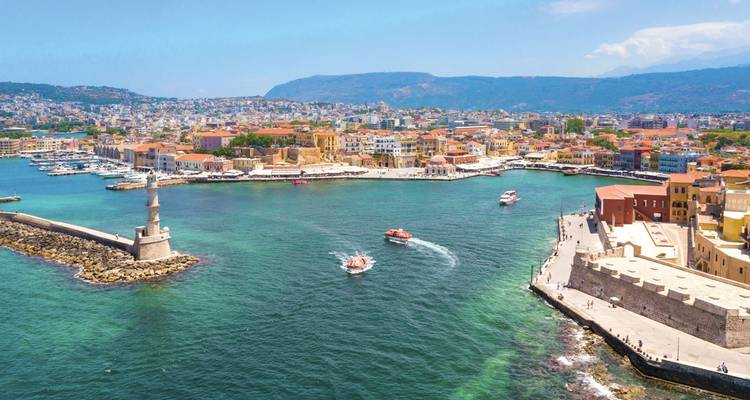 Luchtfoto van mediterrane kuststad met vuurtoren die de haven bewaakt en boten die turkooizen watersporen achterlaten.