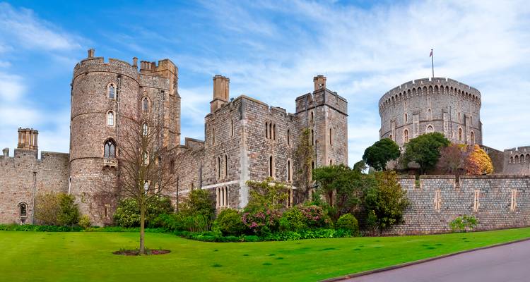 Vue panoramique des tours de pierre grise et des remparts du château de Windsor contre un ciel bleu vif avec des jardins soigneusement entretenus au premier plan.