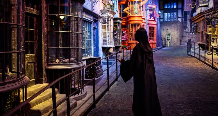 Un visiteur encapé déambule le long des devantures magiques d'une ruelle à la Harry Potter éclairée par des étalages colorés et des fenêtres d'époque.