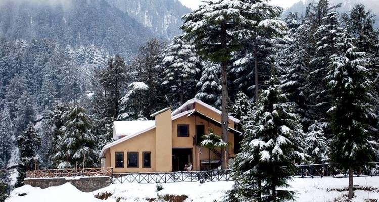 Schneebedeckte Almhütte eingebettet zwischen hohen Tannen mit dahinter aufragenden Bergen