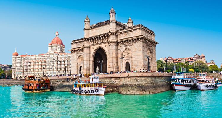 Historische Gateway of India Bögen erheben sich neben türkisfarbenem Hafen mit bunten Touristenbooten und dem Taj Mahal Palace Hotel dahinter