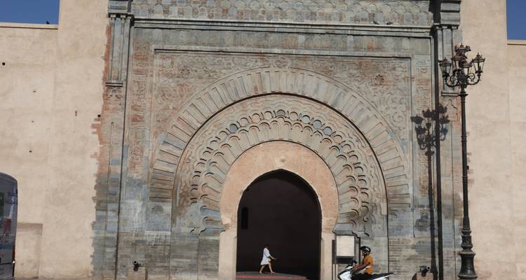 Historisches Bab Agnaou Tor in Marrakesch mit einem einzelnen Fußgänger und vorbeifahrendem Roller bei hellem Tageslicht.