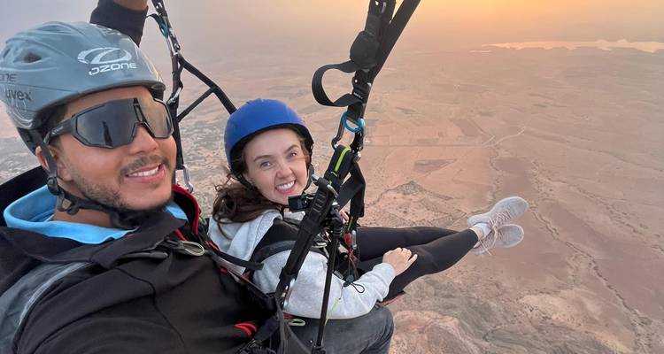 Tandem-Paraglider lächeln in der Luft über einer weitläufigen Wüstenlandschaft bei diesigem Sonnenuntergang.