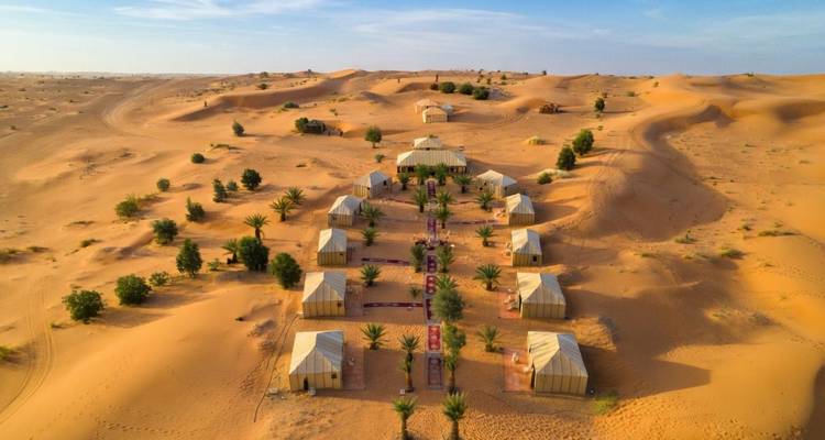 Vue aérienne d'un campement de tentes de luxe disposé en rangées ordonnées au milieu des dunes dorées du Sahara parsemées de palmiers.
