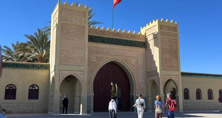 Des visiteurs se rassemblent devant une porte marocaine ornée avec de hauts murs crénelés et un carrelage complexe sous un ciel bleu profond.