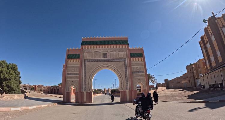 Plan large d'une porte monumentale dans une ville marocaine aride alors que des scooters et des piétons passent en dessous.