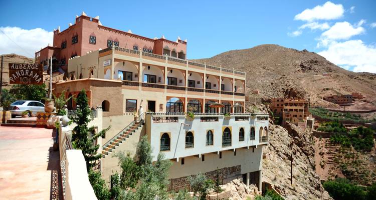 Auberge à flanc de falaise avec terrasses et arcades surplombant une vallée de montagne escarpée sous un ciel bleu.