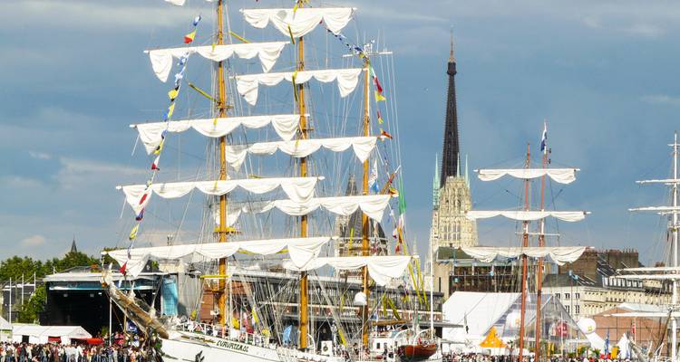 Altos veleros con velas blancas ondulantes atracados a lo largo del Sena durante un festival marítimo en Rouen, con la aguja de la catedral alzándose detrás.