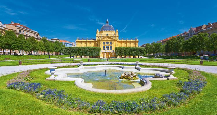 Pavillon d'art jaune néoclassique précédé d'une fontaine et de pelouses soignées sous un ciel éclatant