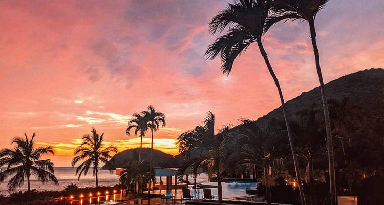 Piscine de complexe tropical et silhouettes de palmiers sur fond de coucher de soleil caribéen orange vif au-dessus de la mer.