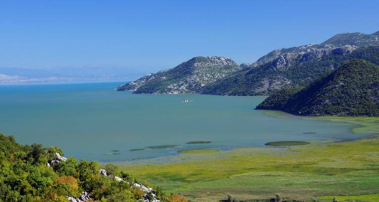 Weitläufiger grün-blauer See, umgeben von bewaldeten Hügeln und fernen Bergen unter einem klaren blauen Himmel