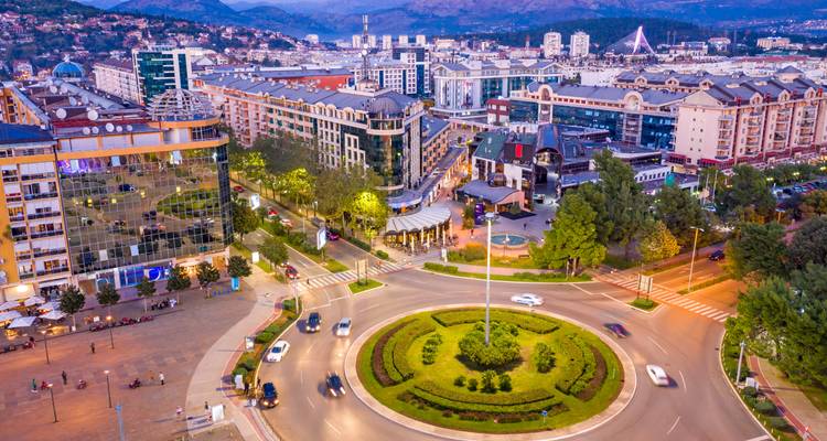 Luftaufnahme einer modernen montenegrinischen Stadt mit einem grünen Kreisverkehr, umliegenden Gebäuden, Autos und fernen Bergen unter klarem Licht.