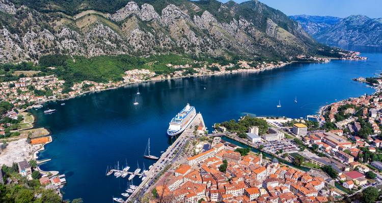 Atemberaubende Luftaufnahme der Bucht von Kotor mit einem Kreuzfahrtschiff, das neben der Altstadt mit roten Dächern anlegt, und dramatischen Bergen, die das tiefblaue Wasser umgeben.