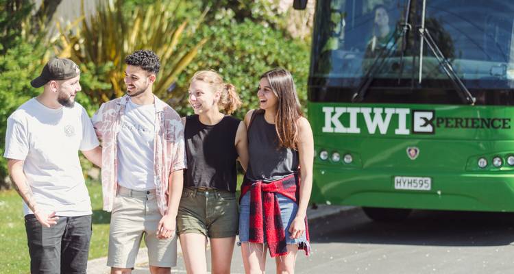 Quatre jeunes voyageurs discutant joyeusement à côté d'un bus de tourisme vert Kiwi Experience.