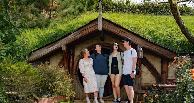 Groupe d'amis souriants à l'extérieur d'une maison de hobbit au toit de chaume entourée de fleurs.