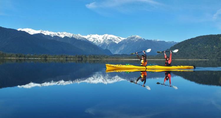 Deux kayakistes levant leurs pagaies sur un lac alpin calme avec des montagnes enneigées se reflétant dans l'eau.
