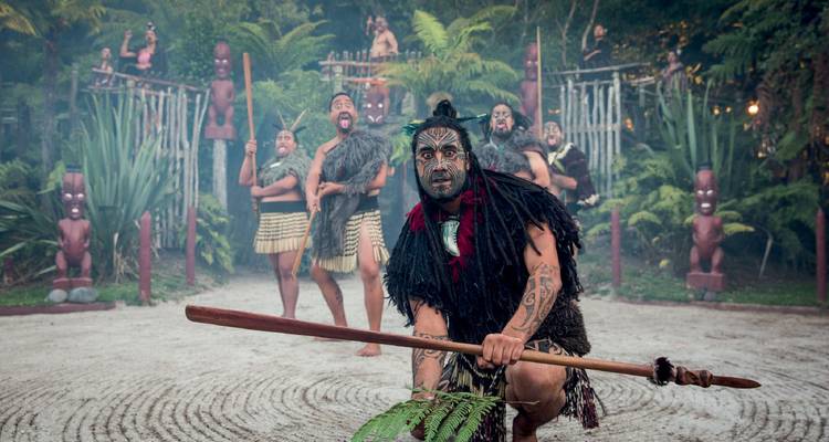 Des artistes maoris en costume traditionnel exécutent un accueil culturel avec des statues en bois sculptées derrière eux.