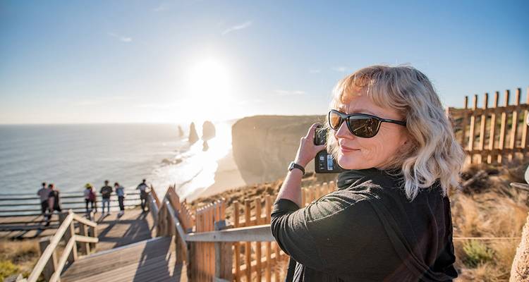 Femme en lunettes de soleil photographiant les pitons rocheux ensoleillés des Douze Apôtres depuis un belvédère en bois le long de la Great Ocean Road.